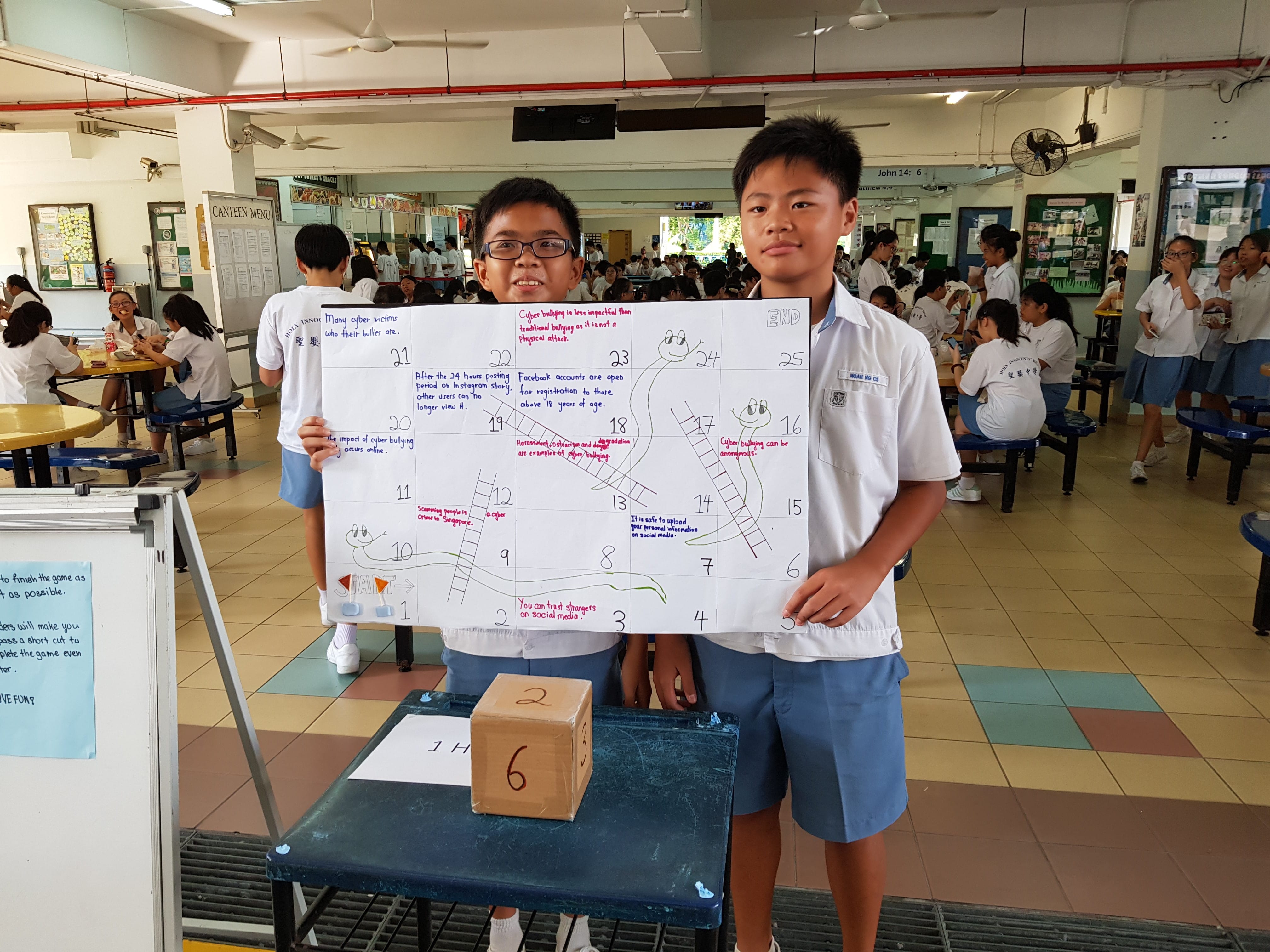 Two school boys in uniforms hold a handmade snakes and ladders board game about cyberbullying in a school canteen.